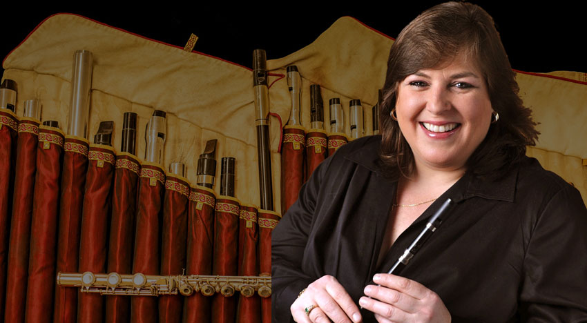 A woman holds a wooden penny whistle as she looks at the camera. She is smiling. The background shows an image of an array of penny whistles in an open cloth carrying case.